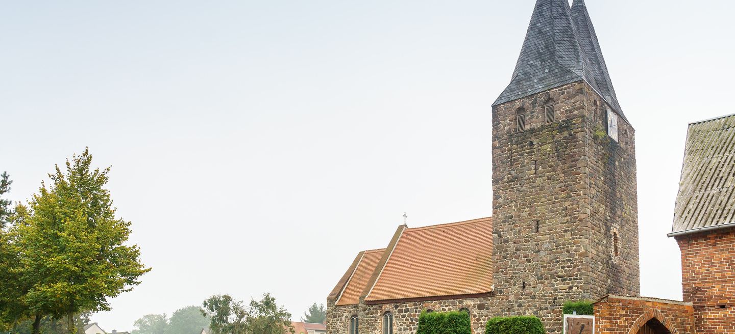 Feldsteinkirche in Klein Schwechten mit wuchtigem Westturm, dunklem Spitzhelm und rotem Ziegeldach über dem Schiff, im Morgennebel der Altmark.