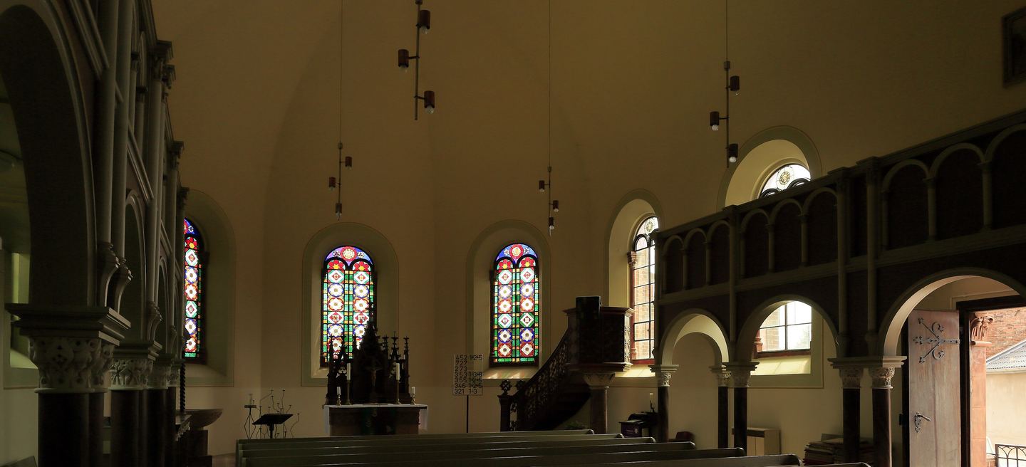 Innenraum der Dorfkirche Großkühnau mit Bankreihen, bunten Glasfenstern, hölzerner Kanzel und der Empore auf schlanken Säulen.