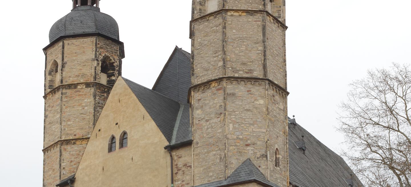 St. Andreas in Lutherstadt Eisleben mit zwei Bruchsteintürmen, hellem Giebel und Schieferdächern unter grauem Winterhimmel, rechts ein kahler Baum.