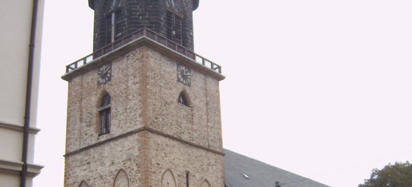 Der Bruchsteinturm der Marienkirche Haldensleben mit zwei Zifferblättern, umlaufender Brüstung und dunkler Haube, rechts das Schieferdach des Kirchenschiffs.