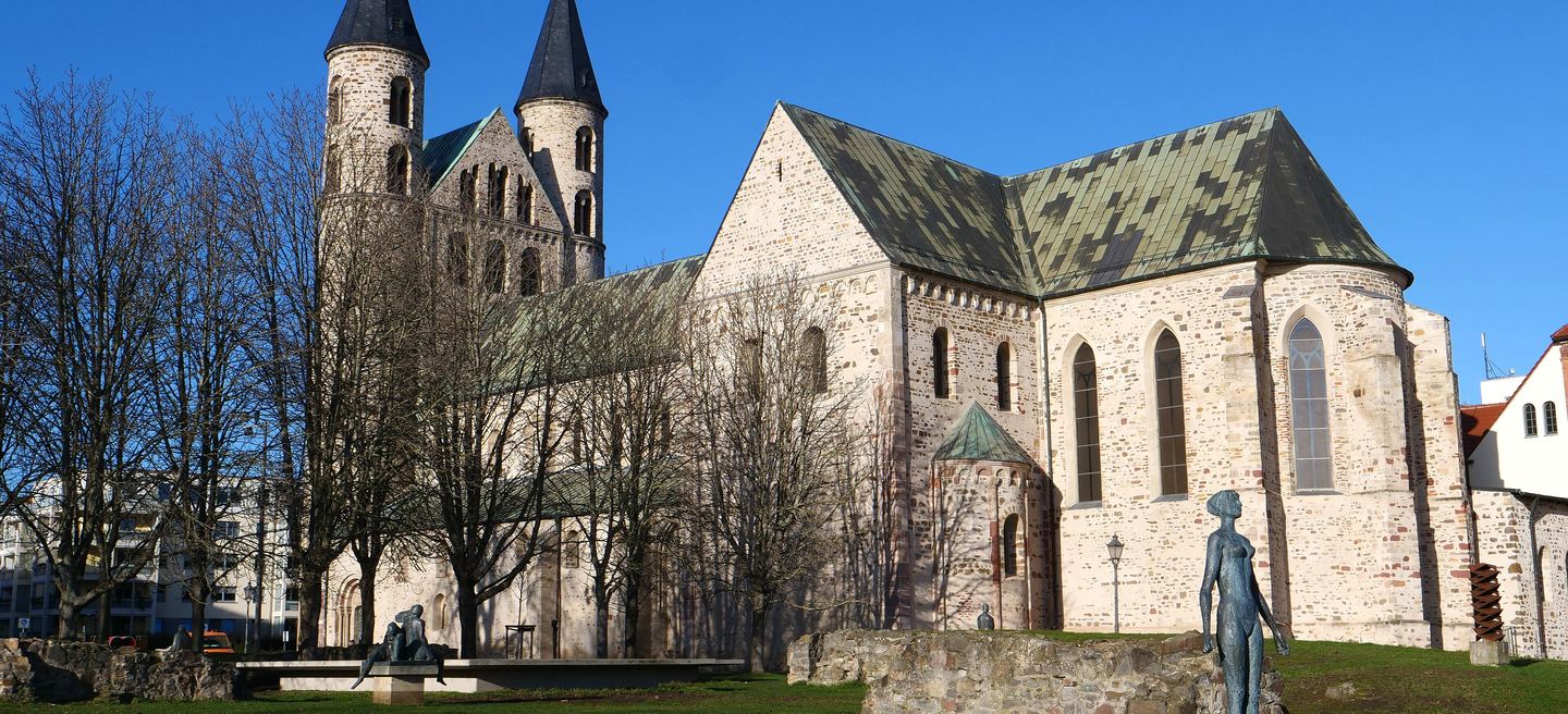 Kloster Unser Lieben Frauen in Magdeburg von Südosten: romanische Doppelturmfassade, gemustertes Kupferdach und Skulpturen im Park vor blauem Himmel.