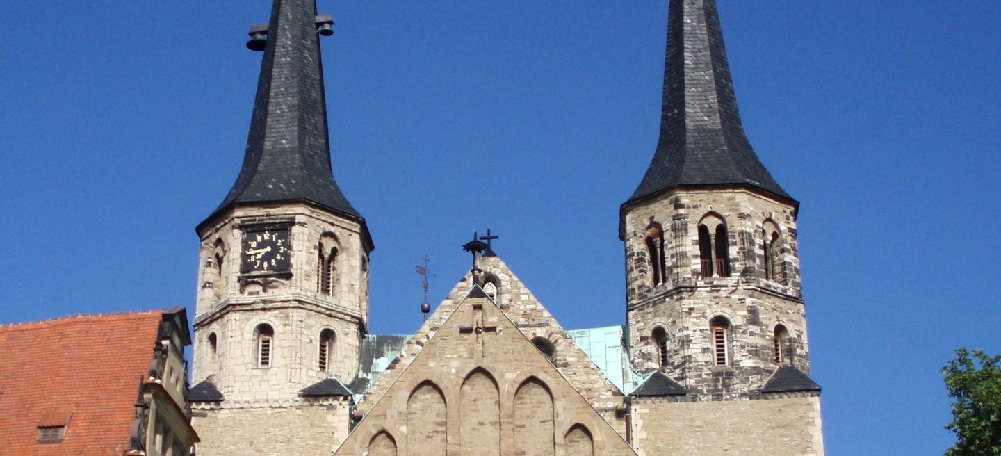 Die beiden Westtürme des Merseburger Doms mit dunklen Turmhelmen und der Giebel des Langhauses vor blauem Himmel, links das Schloss.