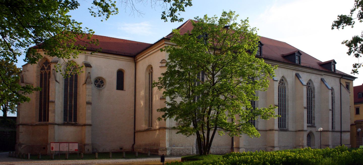 Langgestreckte Südseite des Zeitzer Doms St. Peter und Paul mit hellem Mauerwerk, rotem Ziegeldach, Dachgauben und gotischen Maßwerkfenstern hinter Bäumen.