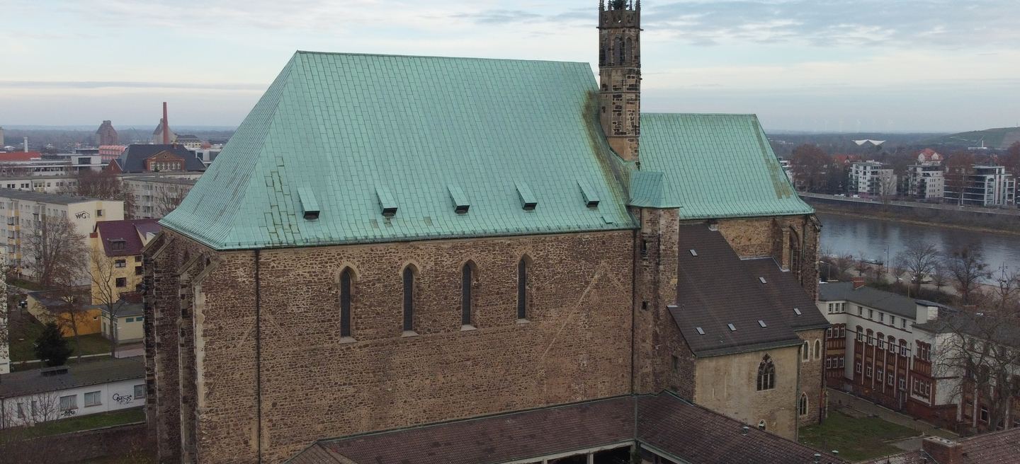 Luftbild der Wallonerkirche in Magdeburg mit grün patiniertem Kupferdach, hohem Bruchsteinschiff und schlankem Turm, dahinter die Elbe und die Stadt.