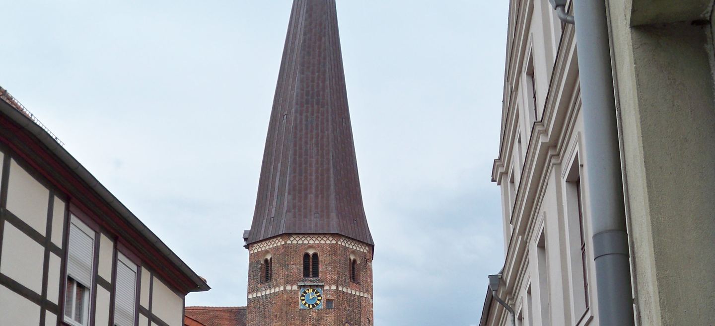 Der hohe Backsteinturm der Marienkirche Salzwedel mit spitzem Helm und Zifferblatt, gesehen aus einer engen Gasse zwischen Fachwerk- und Putzhäusern.