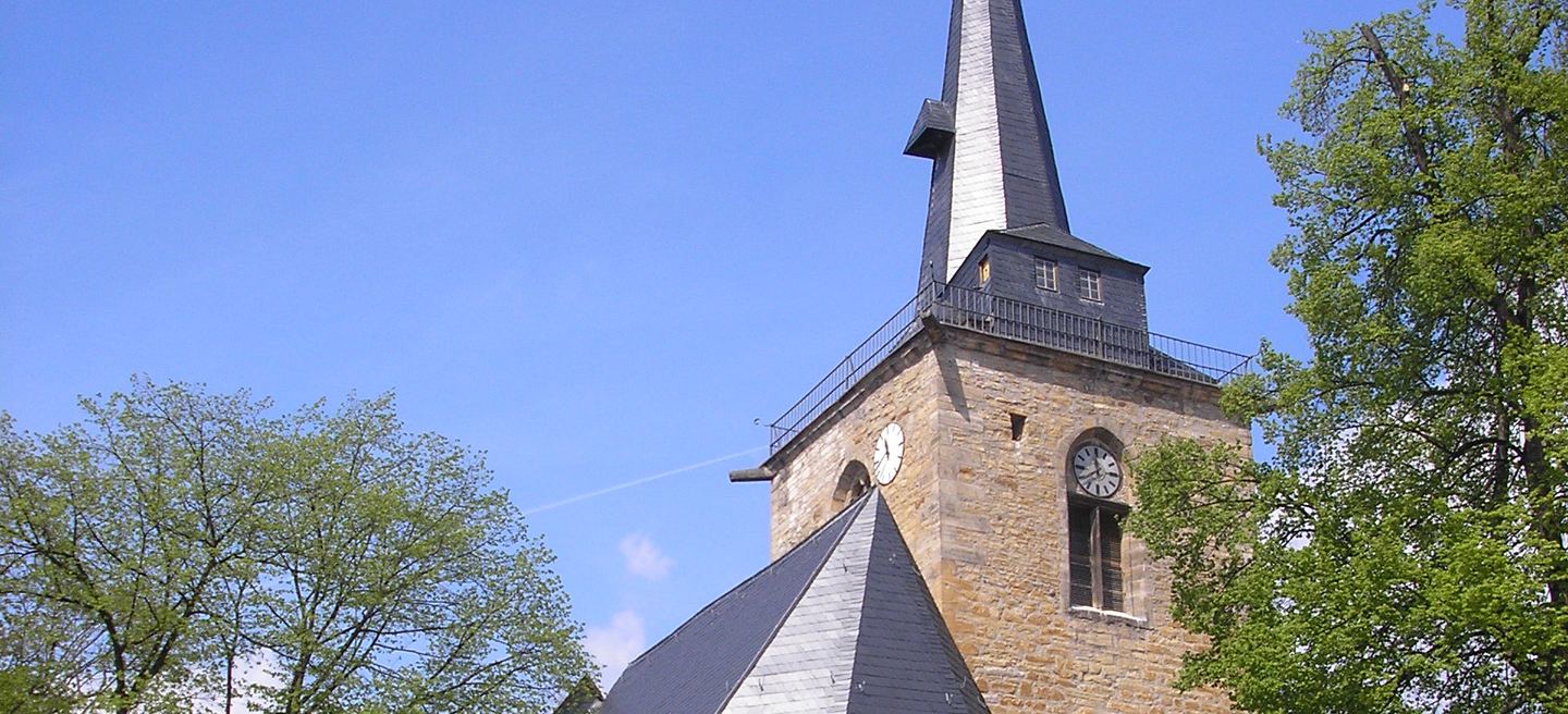 Der Sandsteinturm der Bonifatiuskirche Sömmerda mit Uhr, umlaufendem Umgang und geschwungenem Schieferhelm, daneben das Schieferdach des Langhauses zwischen Bäumen.