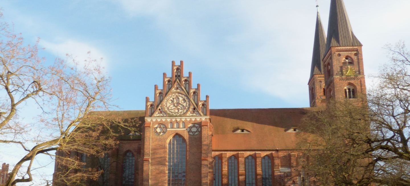 Backsteingotischer Dom St. Nikolaus in Stendal mit Maßwerkgiebel, hohen Chorfenstern und dunkel gedecktem Turmhelm hinter kahlen Bäumen.