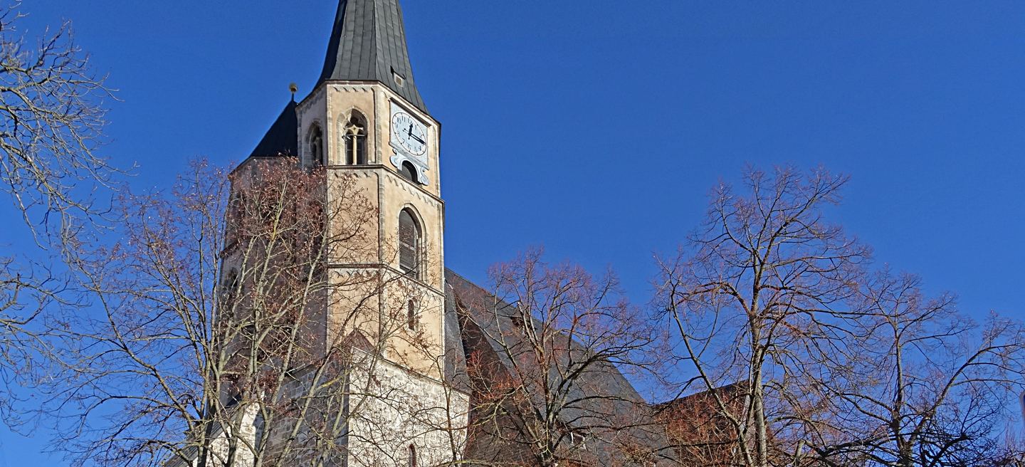 St. Blasii in Nordhausen mit hellem Bruchsteinturm, blauem Zifferblatt und dunklem Helm, umgeben von kahlen Bäumen vor tiefblauem Himmel.