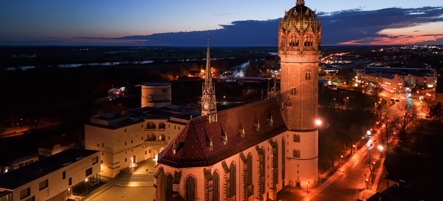 Luftbild der Wittenberger Schlosskirche in der Abenddämmerung: der runde Turm mit goldener Krone, das gemusterte Dach und die beleuchtete Stadt.