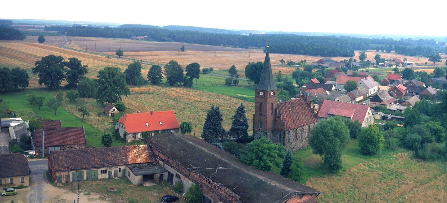 Luftbild des Dorfes Groß-Mehßow mit seiner Backsteinkirche: Turm mit Uhr und Spitzhelm über Gehöften, Scheunen und weiten Feldern.