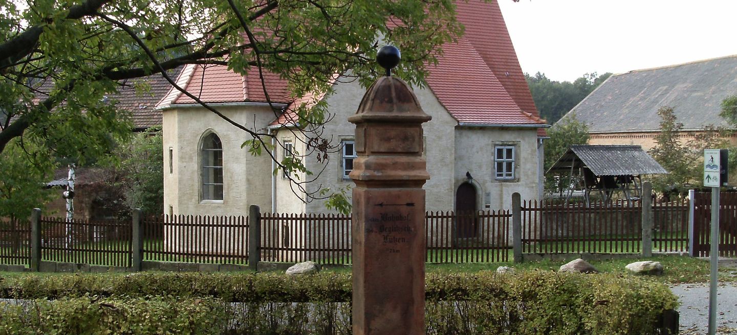 Die Dorfkirche von Dehlitz an der Saale mit verputztem Schiff, rotem Ziegeldach und schmiedeeisernem Zaun, dahinter Gehöfte.