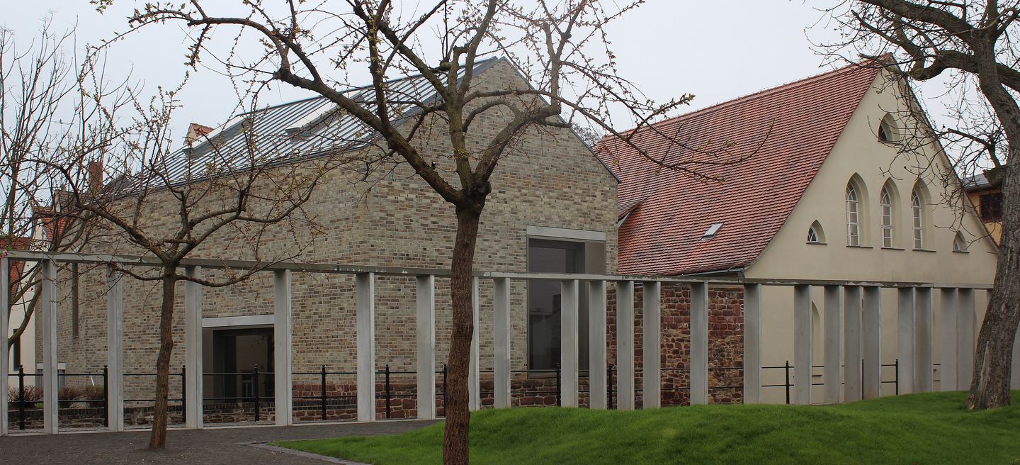 Luthers Geburtshaus in Eisleben von der Rückseite, mit hellem Museumsneubau zwischen Bruchsteinmauern und kahlen Obstbäumen.