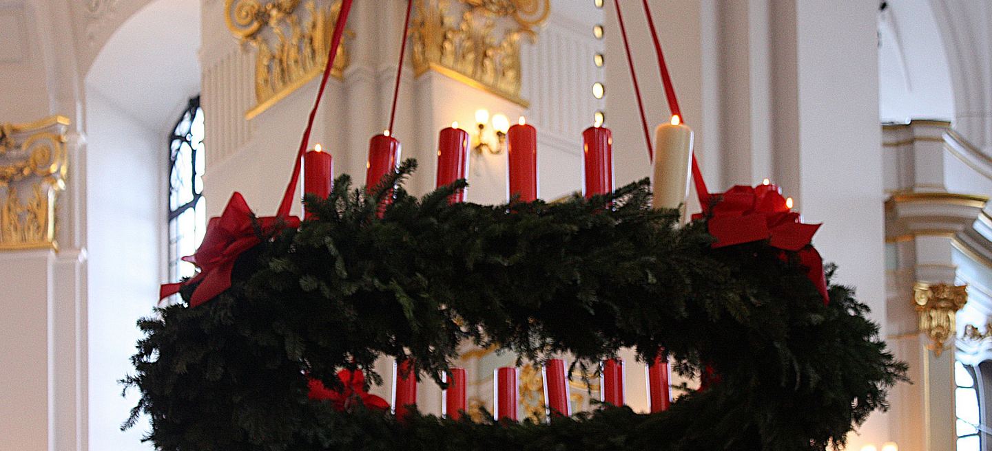 Großer Adventskranz mit roten Kerzen und Schleifen, an Seilen im hellen Kirchenschiff aufgehängt.