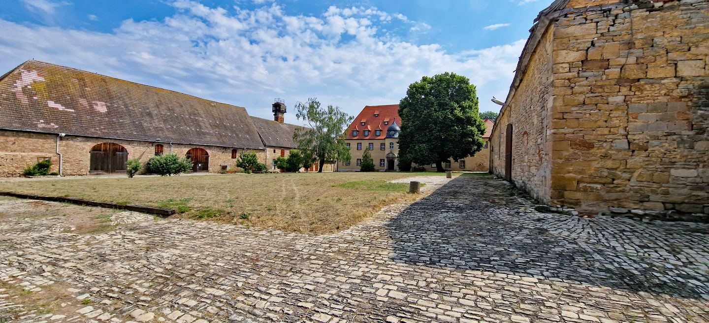 Der Klosterhof von Memleben mit Bruchsteinmauern, Wirtschaftsgebäuden und gepflastertem Weg unter blauem Himmel.