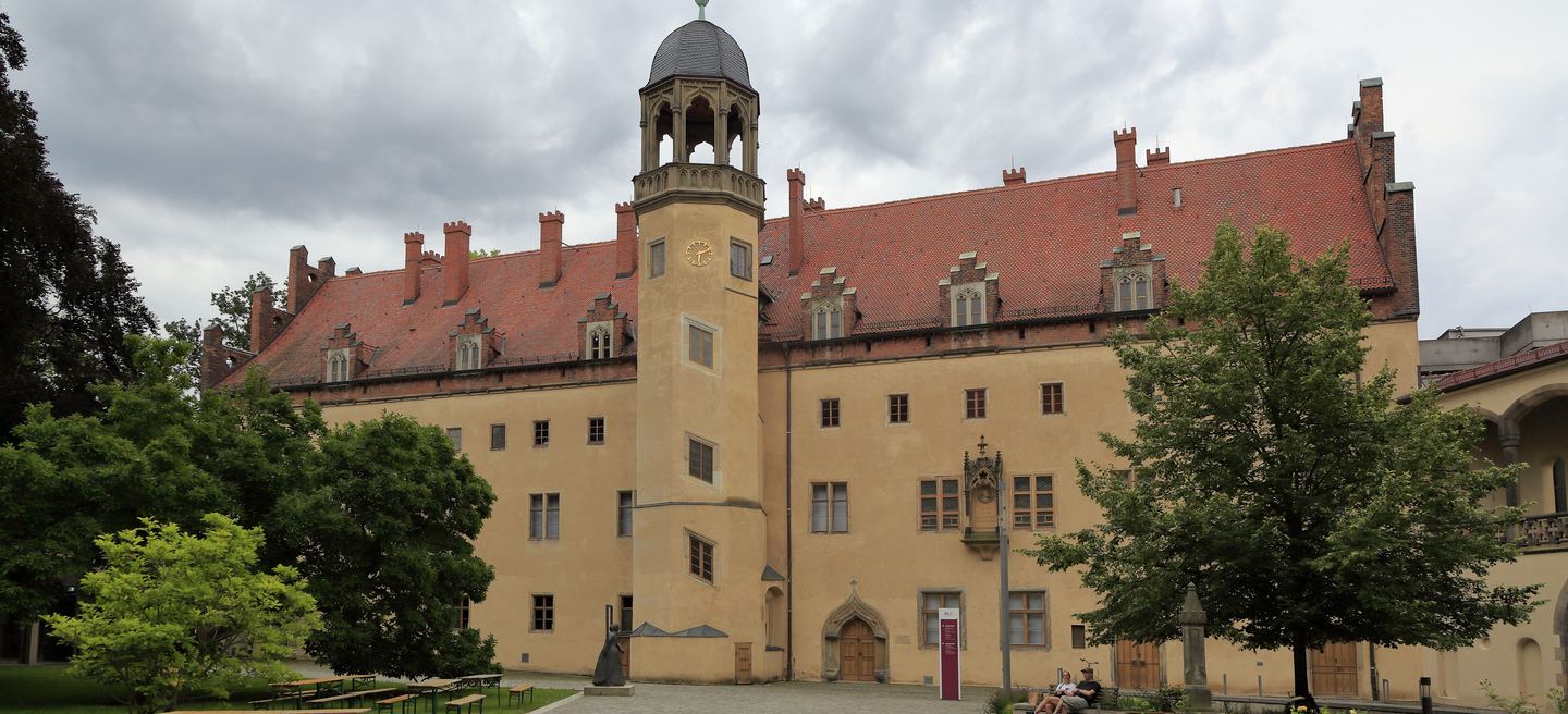 Das Lutherhaus in Wittenberg, ein langgestreckter Renaissancebau mit Treppenturm und rotem Ziegeldach hinter Bäumen.