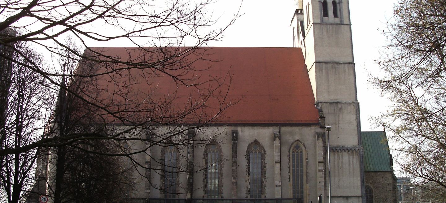Die Magdeburger Johanniskirche mit gotischem Chor, hohen Maßwerkfenstern und grün gedecktem Dach hinter kahlen Zweigen.