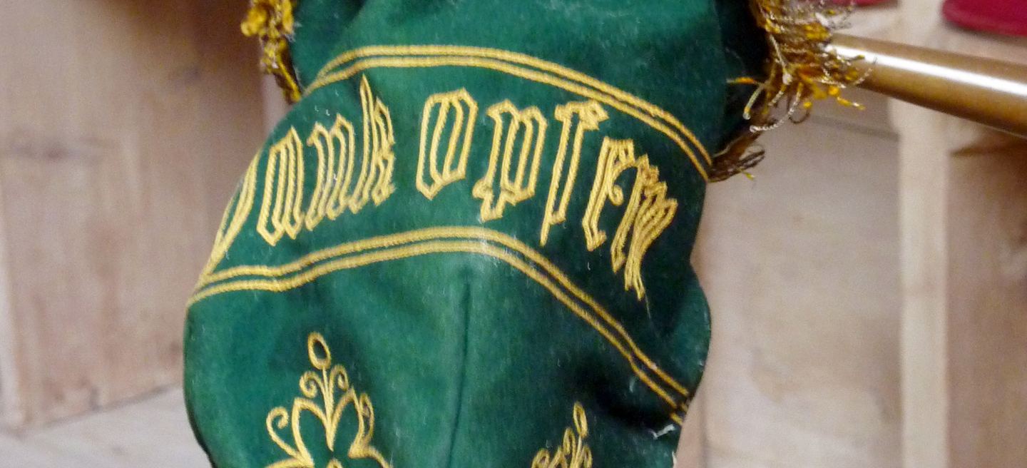 Grüner Klingelbeutel mit goldener Stickerei Dankopfer am hölzernen Stiel, aufgenommen in einer Dorfkirche.